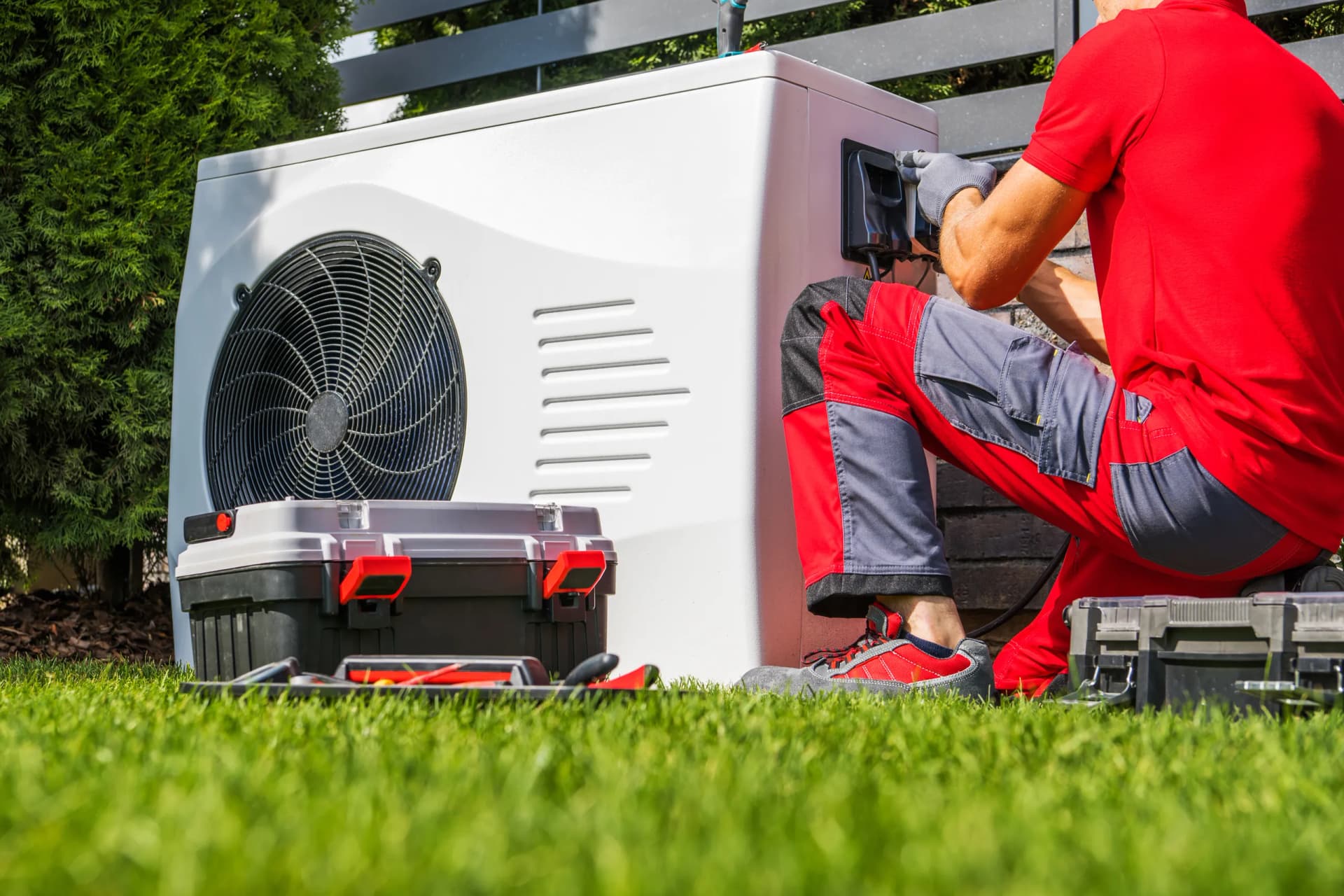A heating engineer in workwear kneels on a lawn, installing or servicing an outdoor heat pump unit.
