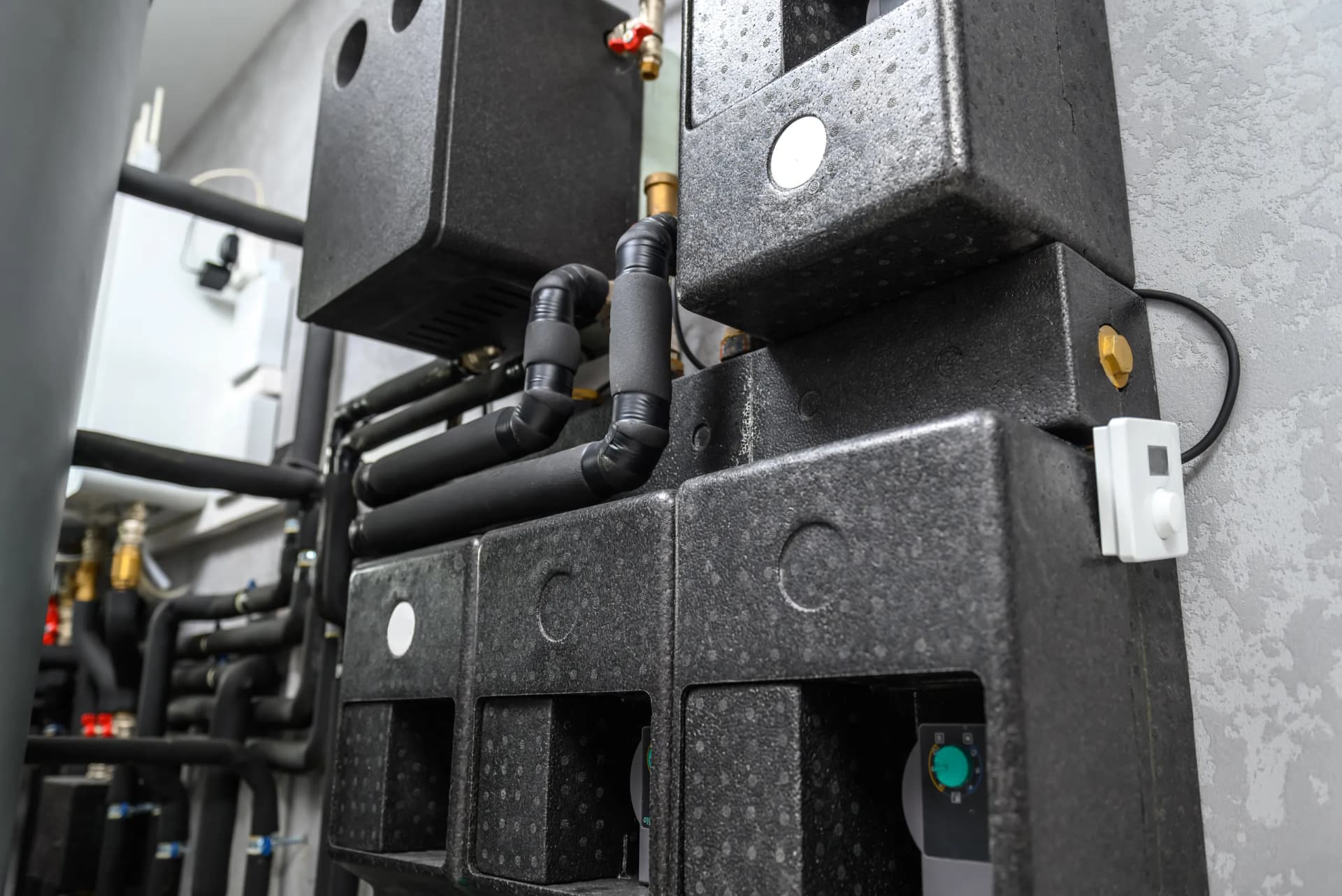 A modern heating system manifold with insulated pipes and grey insulation covers against a textured grey wall.